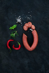 Smoked salami sausage with rosemary, chili pepper, basil and salt over black slate stone background, top view.