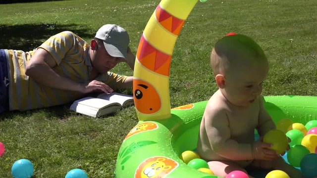 Serious student man dad study book and look after infant baby child sitting in kiddie pool full of colorful balls on garden meadow. Static shot. 4K 
