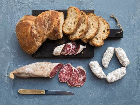 Wine snack set. Italian slami sausages and rustic bread on dark wooden board over a rough grey-blue concrete background