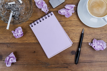 Pink sheets notebook with spiral in the center of the wooden brown table with black pen aside and cup of coffee on top right and glass ashtray with lighted cigarette crumpled paper balls from above