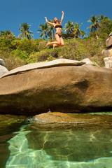 femme qui saute en riant sur des rochers au bord d'une eau cristalline 