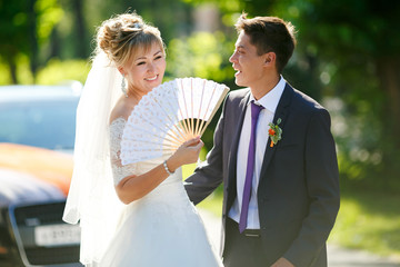 Groom and bride with fan on street illuminated by sun, car in background.