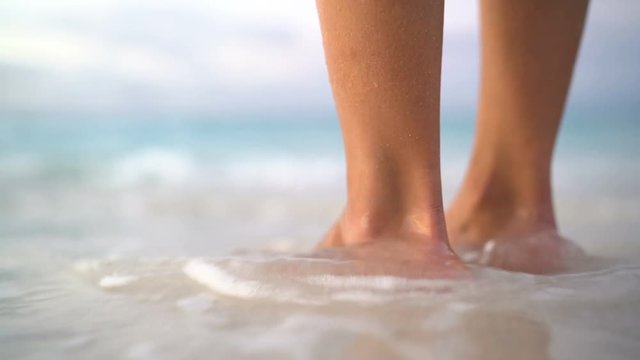 Close up of woman feet on beach. Girl getting her toes wet on vacation. Calm serene relaxing scene on with water splashing on feet in ocean on beautiful beach. 