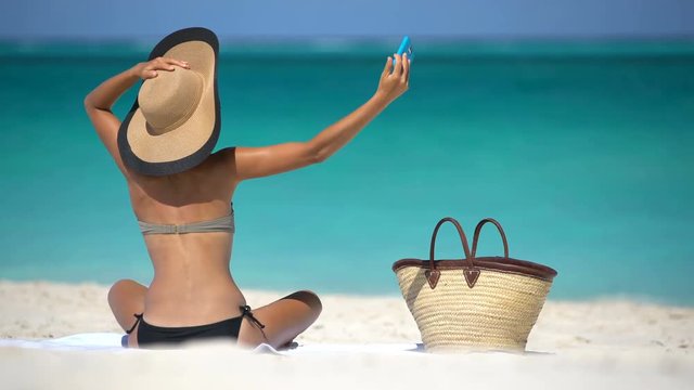 Woman taking selfie while sunbathing on beach. Woman in bikini is photographing through smart phone during vacation. Sexy lady is enjoying summertime at beach in the Caribbean.