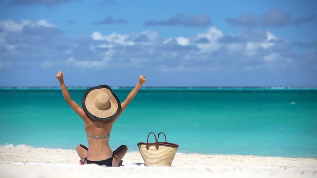 Happy cheering joyful young woman on beach in bikini celebrating with arms raised by beach bag. Pretty female is sunbathing during vacation. Sexy lady is enjoying summertime against turquoise sea.