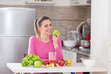 Healthy cheerful woman eating and apple an sitting in kitchen.