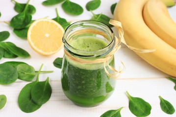 Green spinach and banana smoothie benefits. Green smoothie drink in glass jar, fresh fruit scattered white table. Summery refreshing drink