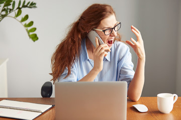 Portrait of an angry businesswoman in glasses shouting on the mobile phone while sitting at desk in...