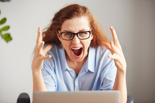 Portrait of a young attractive businesswoman with frustrated look working on laptop at office. Outraged screaming freelance student looking at camera with desperate expression: I hate this computer