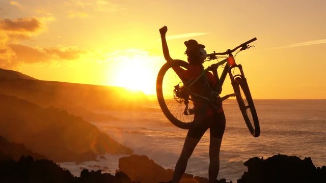 Happy MTB cyclist mountain biking raising arms lifting bike by sea during sunset celebrating successful achievement. Fit woman in sportswear with bicycle. Cheerful lady is enjoying nature on her bike