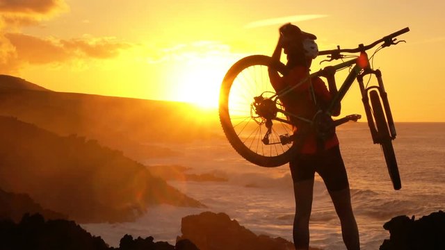 Aspirations. Active lifestyle MTB cyclist mountain biking woman cheering happy raising arms lifting bike by sea during sunset celebrating successful achievement. Person with bicycle in amazing nature.