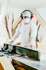 Carpenter working on wood machine in a woodworking factory, wearing face mask and headphones for noise