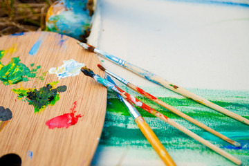 Used brushes near picture with an artist's palette of colorful oil paint for drawing and painting lying on the grass, closeup