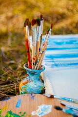 Fototapeta premium Used brushes in the vase near picture with an artist's palette of colorful oil paint for drawing and painting lying on the grass