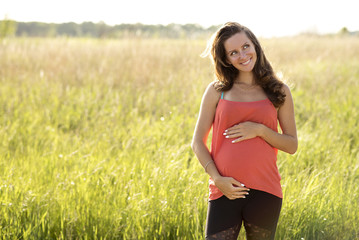 Young beautiful pregnant woman holding tummy smiling, in red a light summer dress, happy on meadow the grass 