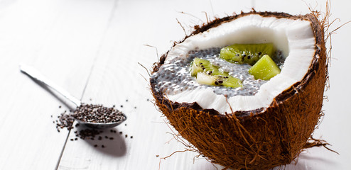 Chia seeds pudding with kiwi fruits in the shell of a coconut on white wooden background.