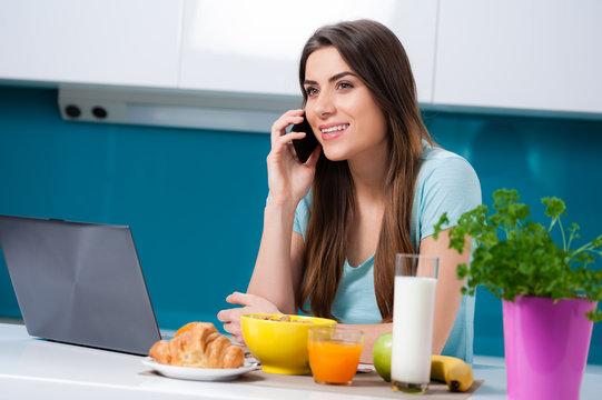 Picture a young woman eating breakfast and taking a phone call, sitting at the kitchen table.