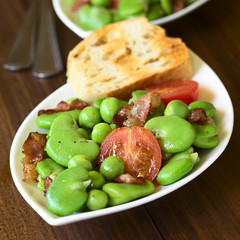 Broad bean, green pea, cherry tomato and fried bacon salad with toasted bread, photographed on dark wood with natural light (Selective Focus, Focus one third into the salad)