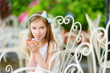 Adorable little girl eating fresh sweet strawberry cake outdoors on warm and sunny summer day