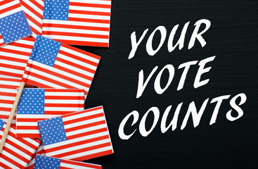 The words Your Vote Counts in white text on a blackboard next to flags of the United States of America as a reminder to vote in the elections