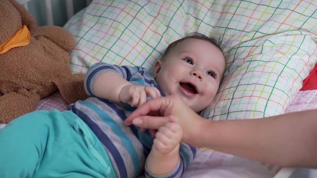 Baby lying in the bed smiling and laughing, mother keeps her hand on baby 
 