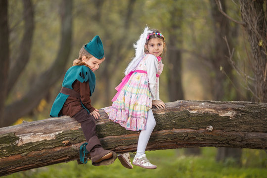 Portrait of a cute little girl dressed up as a fairy sitting on a tree in a forest with a boy in knight costume