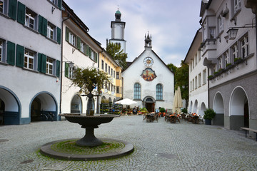 Historic Marktplatz in Feldkirch, Austria, featuring the Johanniterkirche with its unique clock face. Traditional arched buildings and cobblestone streets with medieval architecture.