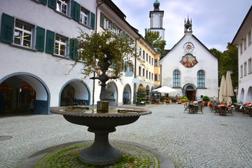 Historic Marktplatz in Feldkirch, Austria, featuring the Johanniterkirche with its unique clock face. Traditional arched buildings and cobblestone streets with medieval architecture.