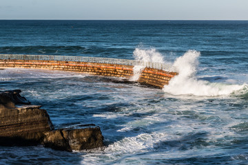 La Jolla Children's pool with nearby rocks and waves crashing over seawall. 