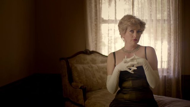 Attractive blonde woman wearing a classic vintage fifties dress and white silk opera gloves, sitting on a chaise lounge.  Originally recorded in 4K and tinted.