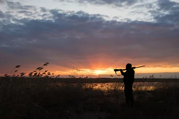 Fotobehang Jacht Silhouet van de jager met het jachtgeweer op een zonsondergangachtergrond