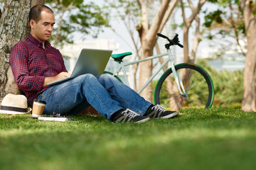 Mixed-race copywriter sitting at tree and working on laptop