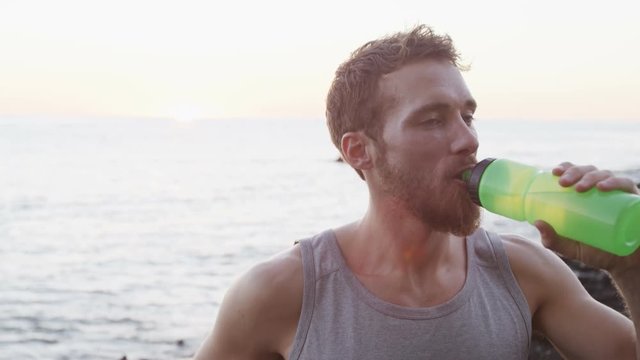 Fitness man drinking water after running workout outside on beach. Thirsty male athlete having cold refreshment drink sweating after intense exercise outdoors. RED EPIC SLOW MOTION.