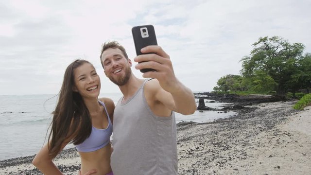 Couple taking selfie after running workout fitness. Good looking young man and woman taking a self-portrait photo with smart phone camera after exercising on a beach. Mixed race couple. RED EPIC.
