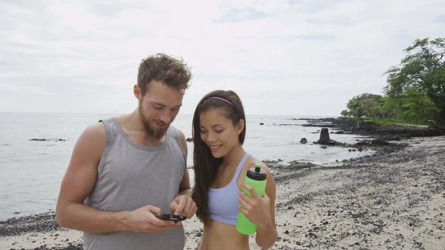 Couple looking at phone app after running workout fitness. Good looking young man and woman using smartphone relaxing after exercising on a beach. Mixed race couple. RED EPIC.