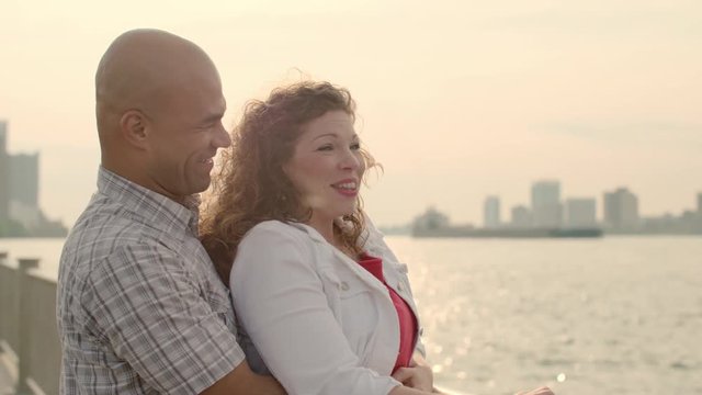 Happy couple hug as they talk and laugh next to the Detroit River. Large ship passes behind them.  Detroit and Windsor, Ontario, skylines visible in background.  Tinted clip.  Slow motion
