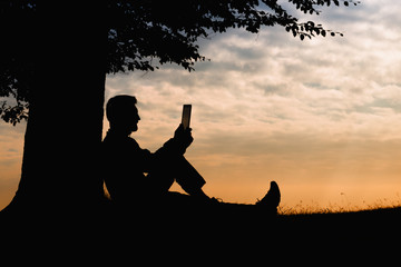 Man silhouette sitting under tree with book on cloudy day outdoor