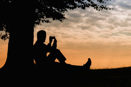 Man silhouette sitting under tree with phone on cloudy day outdoor