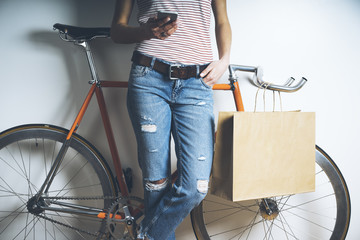 Hipster girl with vintage orange bicycle using modern smart phone, mock-up of blank paper shopping bag with handles