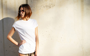 Lovely young girl wearing in a white blank t-shirt and sunglasses posing against a background of a concrete wall in the rays of the setting sun