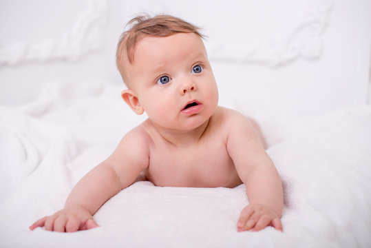 A cute little naked baby is looking into the camera on the white wall.