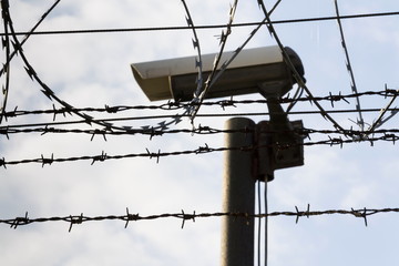 Security camera behind barbed wire fence stretched around prison walls