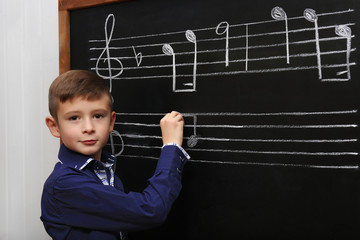 Cute boy writing at the blackboard with musical notes, in the classroom