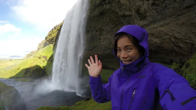 Woman tourist waving hello jacket by majestic waterfall. Happy girl visiting spectacular natural landmark during vacation, the famous attraction. Seljalandsfoss waterfall, Iceland. ACTION CAMERA.