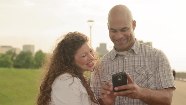 Laughing and smiling couple use a smart phone with Detroit city skyline visible in background.  Slow motion, recorded hand-held at 60fps in 4K.
