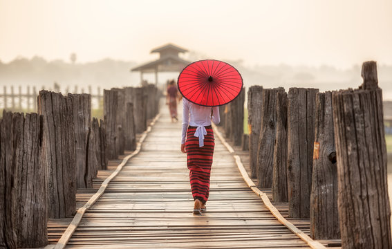 Burmese woman holding traditional red umbrella and walking on U Bein Bridge