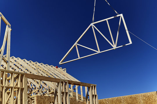A wooden roof truss being lifted by a crane in the air for placement on the roof of a new home