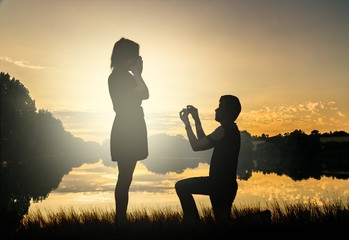 Wedding proposal concept. Young couple have dating at sun set. Man in love is kneeling on knees and giving wedding ring to surprised woman. - obrazy, fototapety, plakaty