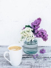 Black coffee in white porcelain cup on vintage wooden table with with branches of lilac in transparent glass vase 