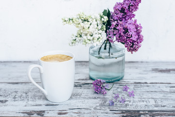 Black coffee in white porcelain cup on vintage wooden table with with branches of lilac in transparent glass vase 